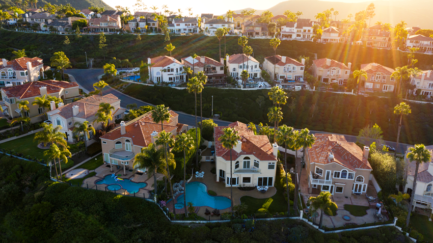 Aerial photograph of a hillside residential neighborhood in Orange County, California at golden hour. Depicts characteristic OC architecture: red-tile Mediterranean roofs, mature palm trees, terraced hillside lots, and backyard pools. Featured image for the 2026 Orange County relocation guide by REALTOR Debbie Sagorin.